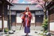 A woman in traditional attire stands in front of a Japanese-style house with cherry blossoms.