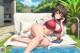 A woman in a red bikini sitting on a white couch next to a pool.